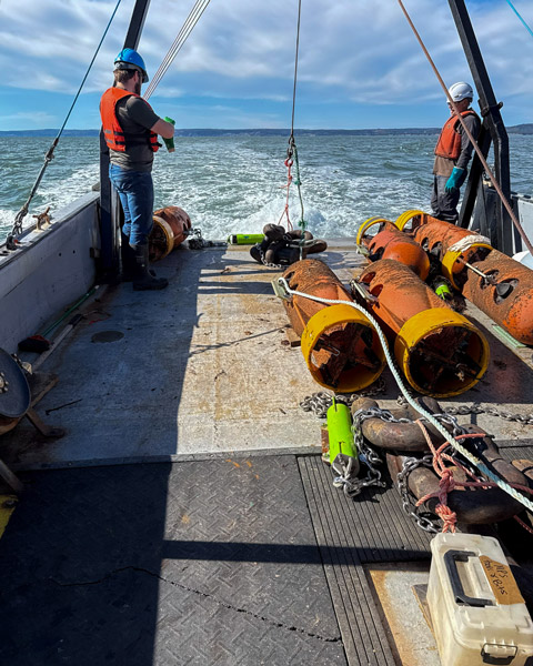 Large orange floats near the back of a ship with two technicians preparing the moorings for launch. 